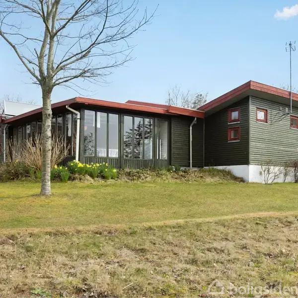 Huset er en moderne, grøn bygning med store vinduespartier, placeret på en græsplæne. Omkring huset står træer og buske. Himlen er blå med spredte skyer.