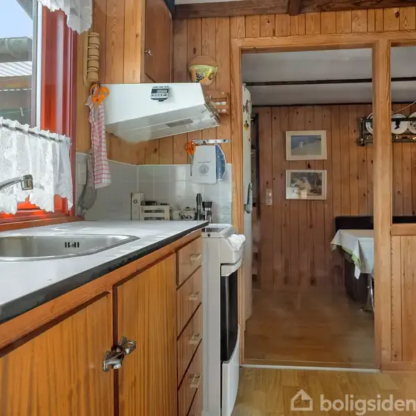 En lille køkken med træpaneler dominerer rummet, hvor en vask og komfur er placeret ved et vindue med blondelangegardiner. I baggrunden ses en åben dør til en stue.