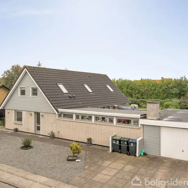 Et parcelhus med skråt tag og flere ovenlysvinduer står ved siden af en garage i et villakvarter. Foran huset er en grusbelagt have med små buske.
