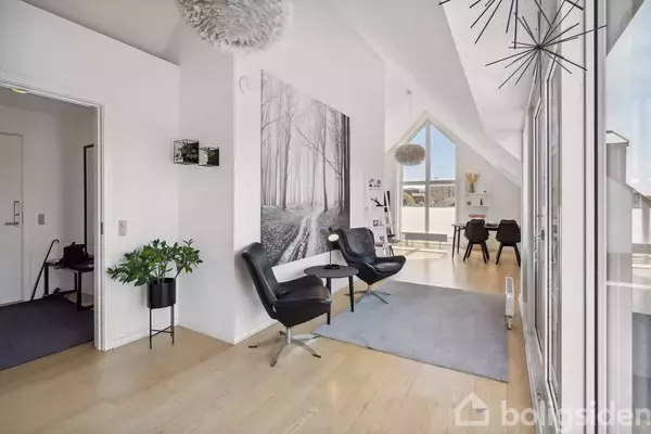 Stue med to sorte lænestole og et lille bord på et lysegråt tæppe. Store vinduer giver udsigt til lys balkon. Vægkunst forestiller en skov i sort/hvid.