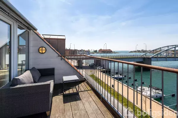 En altan med sofa og bord vender ud mod en bro og vandet. Vandet strækker sig mod en fjern bymæssig baggrund under en klar blå himmel.