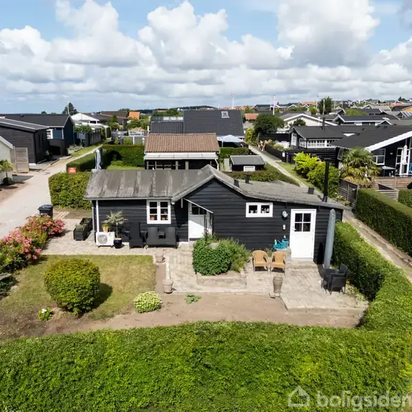 Sort træhus står i en velplejet have med hække og buske, beliggende i et hyggeligt sommerhusområde med flere lignende huse på en solrig dag.