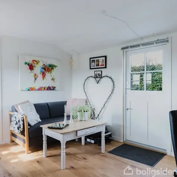 En stue med en sort sofa ved siden af et hvidt kaffebord, dekoreret med en plante og udstilling. Et verdenskort og hjertepynt hænger på væggene. En sort stol ved skrivebordet.
