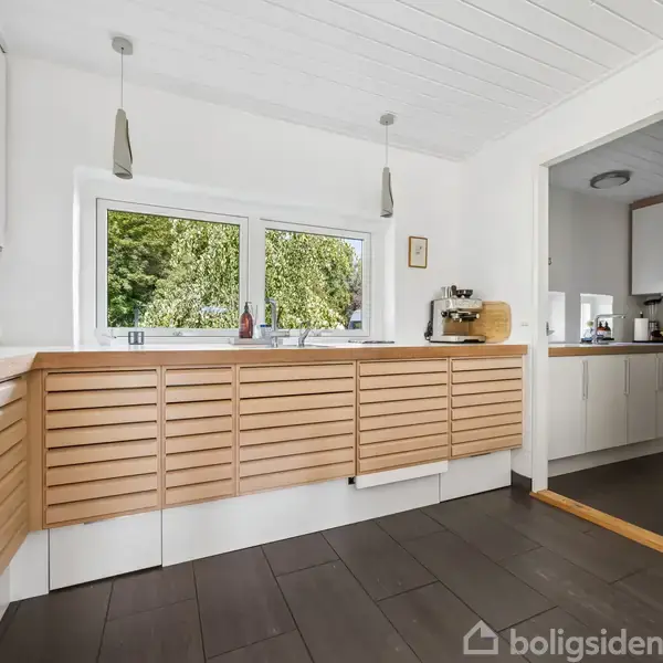 Køkken med lyse træfronter på skabene, bordplade med køkkenmaskiner og en stor vindue. Rummet er lyst og moderne med hængende lamper over bordpladen.