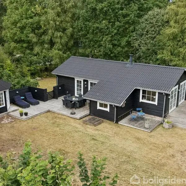 Et sort træhus med terrasse omgivet af græsplæne og træer. Der er havemøbler og liggestole på terrassen. Et lille anneks ses ved siden af huset.
