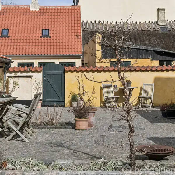 Træmøbler står i en brostensbelagt gårdhave med krukker og planter. Høj, gul mur med en grøn dør skaber en afgrænsning, mens røde tegltage ses i baggrunden under en blå himmel.