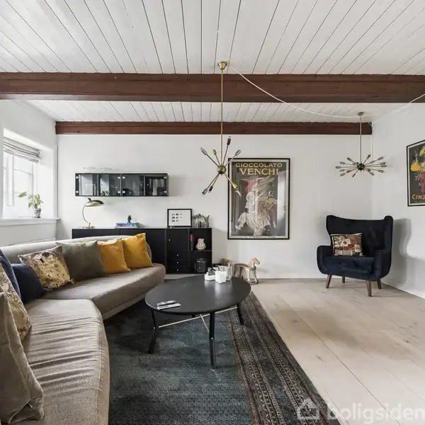 En stue med en grå, buet sofa fyldt med farverige puder står på et tæppe. En mørk lænestol med pyntepuder placeret ved siden af kunstplakater på væggen.