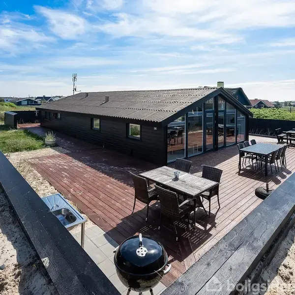 Sort sommerhus med store vinduer står på en hævet træterrasse. To sæt havemøbler og en grill er placeret udenfor. Omgivet af klitter og spredte huse under en blå himmel.