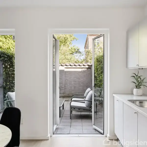 Døre åbnes mod en terrasse med to stole. Indendørs ses et hvidt køkken med vask og grønne planter. Udenfor er der en mur og beplantning.