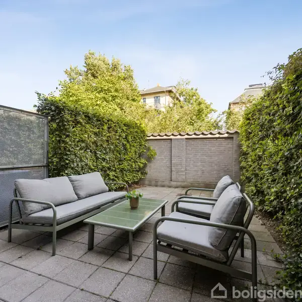 Terrassemøbler står stille på en flisebelagt terrasse, omkranset af grønne buske og en mur, under en blå himmel.