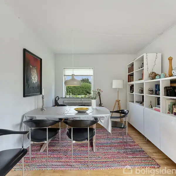 Langbord med hvid dug omgivet af sorte stole står på farverigt tæppe. Vægreol fyldt med bøger og dekorationer står ved siden af. Vindue viser udsigt til grøn have.