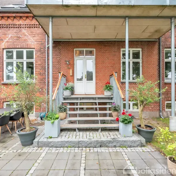 Murstenshus med trappe dekoreret med planter fører op til hvid dør. Omkringliggende flisebelagt gårdhave har havemøbler og krukker. Græsplæne ses i højre side.