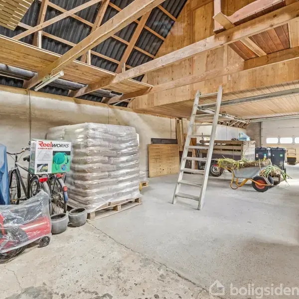 En garage med en træstige, stakke af sække og presenningsovertrukne genstande. En cykel og værktøj er opbevaret langs væggen. Garageporten er lukket i en lys, rummelig struktur.