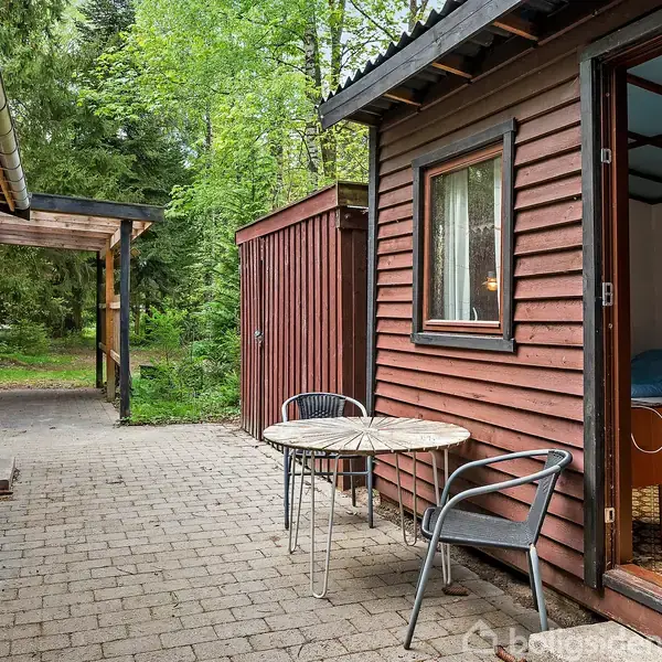 En bord og to stole står på en terrasse af fliser ved siden af en træbeklædt bygning. Dør fører ind i huset. Grønne træer omgiver området i baggrunden.