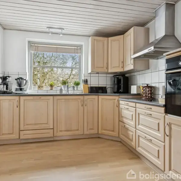 Køkken med lyse træskabe, der står ubrugte, i et rum med hvidmalede vægge og trægulv. Et stort vindue giver udsigt til træer udenfor. Ovne og køkkenredskaber er synlige.