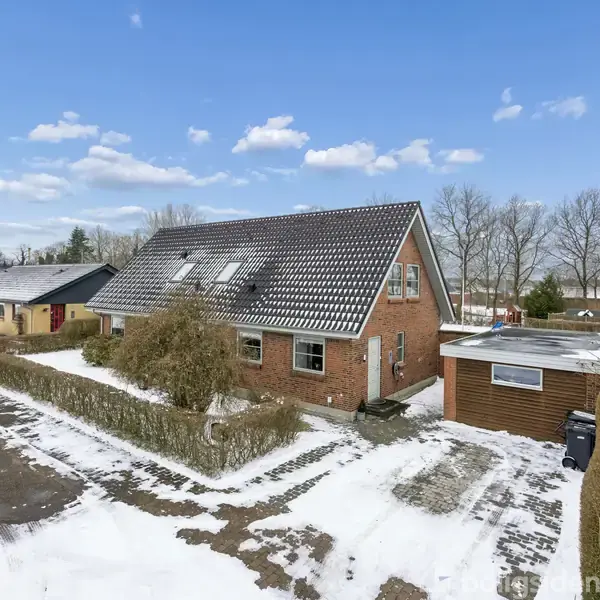 Murstenshus med skråt tag står stille i et snedækket område, omgivet af hække og træer, med flere huse og garager i baggrunden under en blå himmel med få skyer.