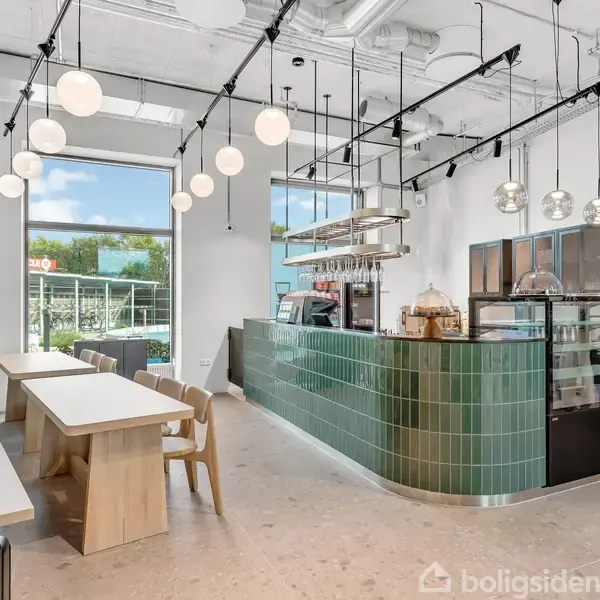 Caféområde med grøn flisedisk, lyskugler hængende fra loftet, store vinduer med udsigt til udendørsområde. Træborde og stole langs væg. Lyse vægge og gulv skaber en moderne atmosfære.