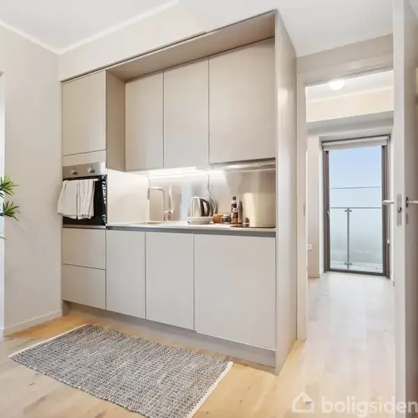 Et kompakt køkken med indbyggede skabe og apparater placeret mod en væg. En grå måtte ligger på det lyse trægulv. Ved siden af står en grøn plante. I baggrunden ses en åben dør mod en balkon med glasværn.