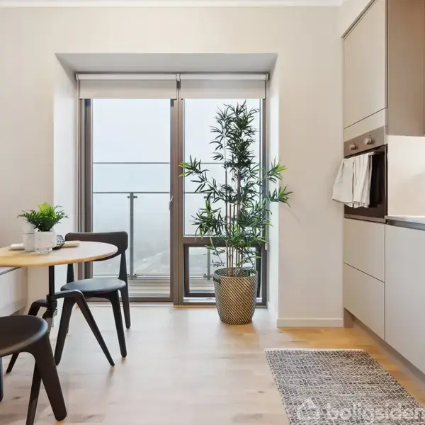 Et moderne køkken med to sorte stole og et rundt spisebord. Der er en stor plante ved en glasdør, og køkkenelementerne er stilfulde med integrerede apparater. Atmosfæren er lys og minimalistisk.