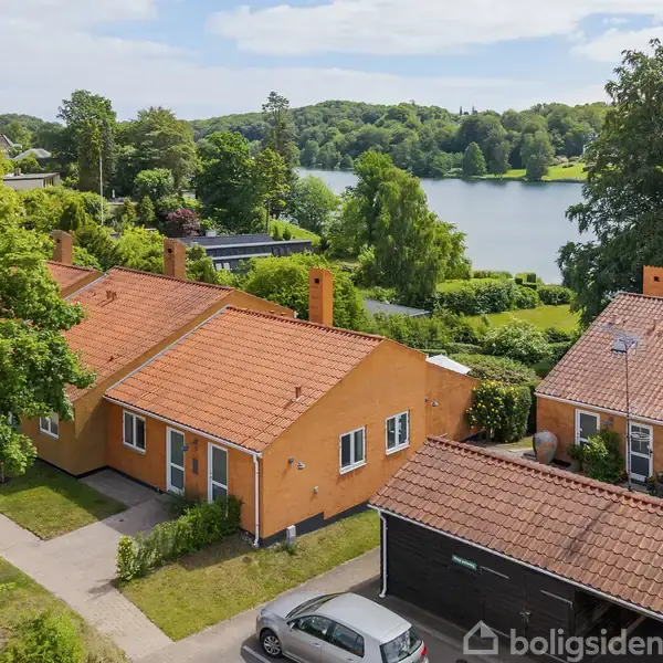 En orangemalet bungalow med teglstenstag ligger ved en sø omgivet af grønne træer. En bil er parkeret foran huset, og andre lignende huse ses i nærheden.