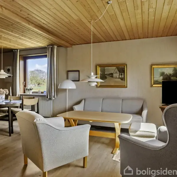 Stue med træloft og lyst trægulv. Sofa og lænestole står omkring et sofabord. Spisebord ved vinduet. Væggene har kunstmalerier, og et fjernsyn står i hjørnet.