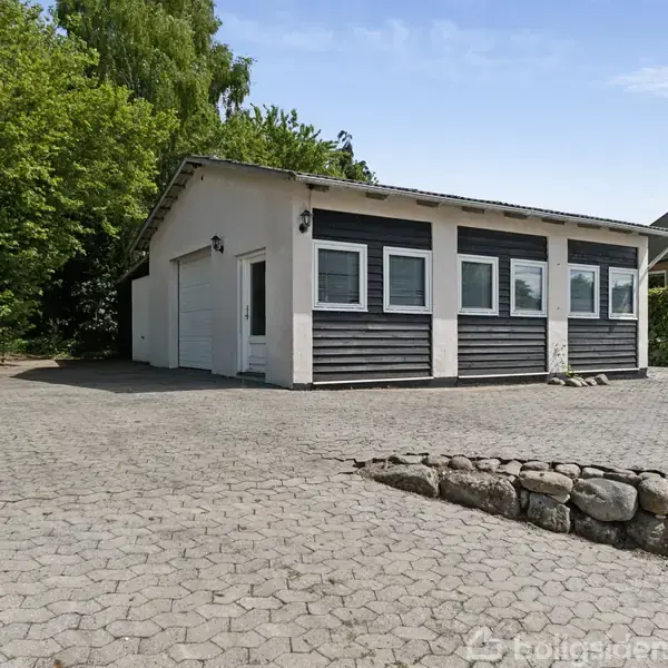 Garagebygning med hvid facade og sorte træpaneler, står i en stenbelagt gårdsplads omgivet af buske og træer, ved siden af en grå murstensmur og solrig himmel.