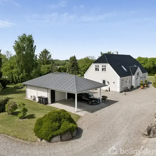 Hvidt hus med sort tag står i en grøn have. En carport ved siden af rummer en bil. Grusvej fører til bygningen, omgivet af buske og græsplæne.