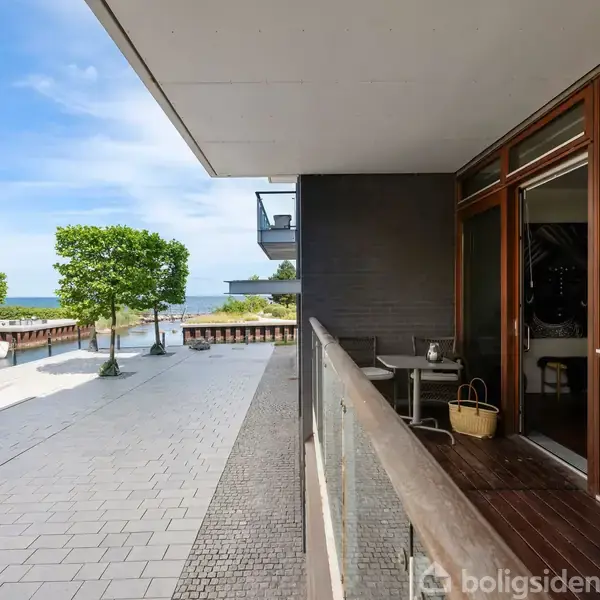 En altan på en moderne bygning med glasrækværk. Ved siden af er en flisebelagt promenade med træer. I baggrunden ses en kystlinje og vandet.