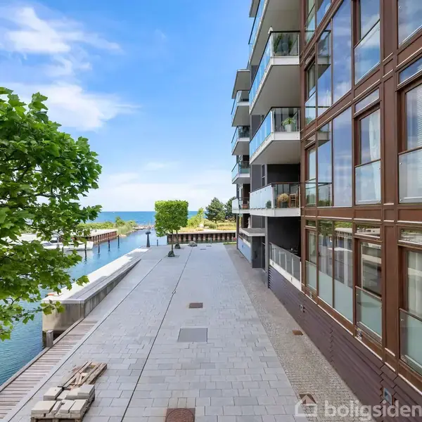 En moderne lejlighedsbygning med altaner står ved en havnefront. Fliser dækker stien ved siden af bygningen, og træer grænser op til vandet under en blå himmel.