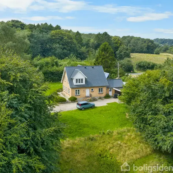 Et hus ligger midt i en frodig, skovklædt landskab. En bil er parkeret foran huset på en grusvej. Omkring det ses grønne træer og marker i baggrunden.