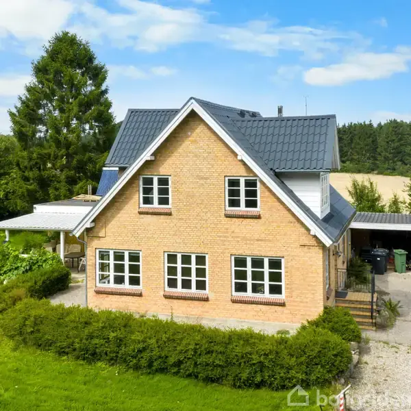 Et enfamiliehus i mursten står stille omgivet af en grøn græsplæne og træer. Til venstre ses en overdækket terrasse, mens en garage ses til højre.