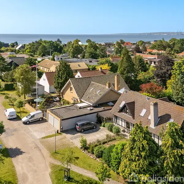 Huse med skrå tage står langs en stille vej i et frodigt boligområde. I baggrunden ses havet. Biler er parkeret foran husene, omkranset af grønne haver og træer.