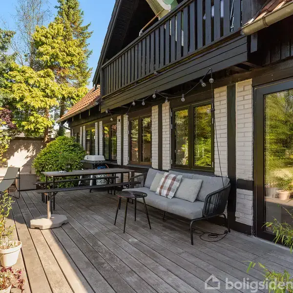 En træterrasse med en sofa og bord i naturlige omgivelser mod en husfacade. Små planter i krukker og græs omkranser terrassen, der vender ud mod en grøn have.