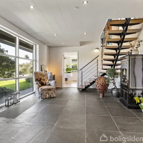 En moderne stue med en åben trappe fører til første sal. Rummet er lyst, med store vinduer, en lænestol, planter og marmorfliser. Udsigt til en grøn have udenfor.