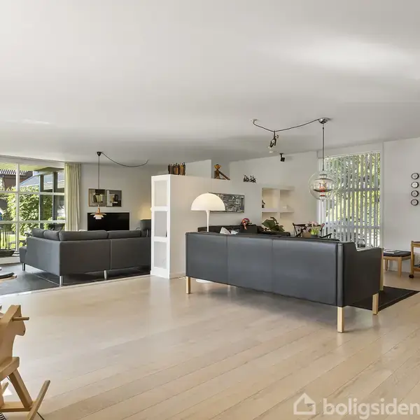 Lædersofaer står i en rummelig stue med lyst trægulv og store vinduer ud til en have. Rummet er indrettet med moderne møbler og dekorative elementer.