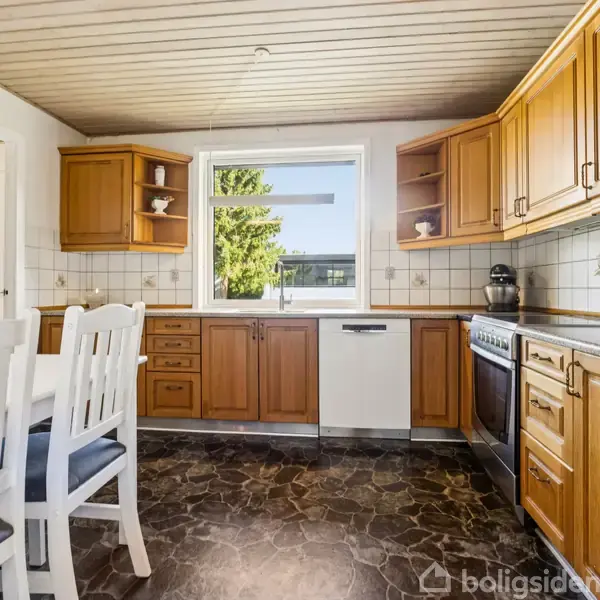 Et køkken med træskabe omslutter et komfur og en opvaskemaskine. Bordet med hvide træstole står foran. Et vindue giver udsigt til et træ udenfor.