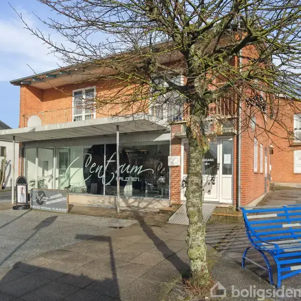 En rød murstensbygning med et skilt, hvor der står "Centrum". Foran bygningen står et træ og en blå bænk. Til venstre er et skilt med "P" for parkering.