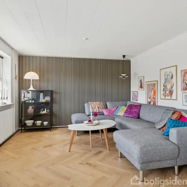 Gråt hjørnesofa står i stue med træparketgulv, farverige puder, sofabord og lampeskab. Væggene er dekoreret med kunstværker. Store vinduer giver naturligt lys.