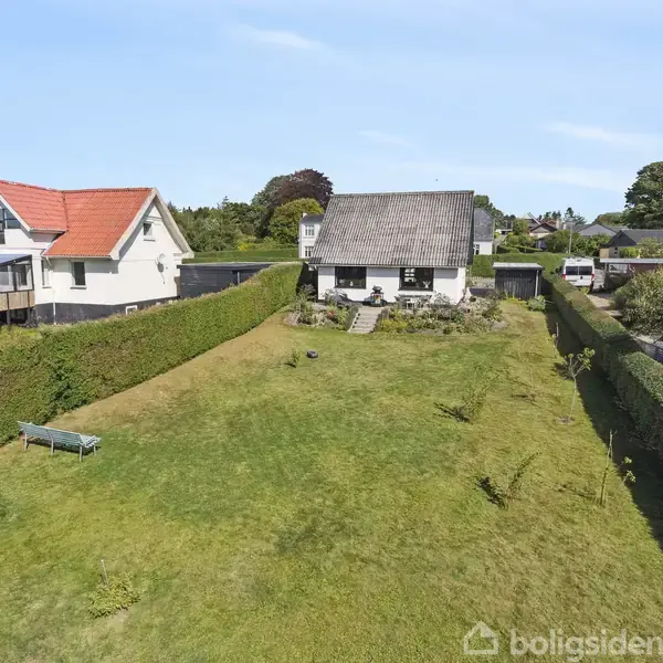 To huse står på en stor, velplejet græsplæne omgivet af hække. Ét hus har et rødt tag og en udendørs terrasse. Solrig himmel i baggrunden.