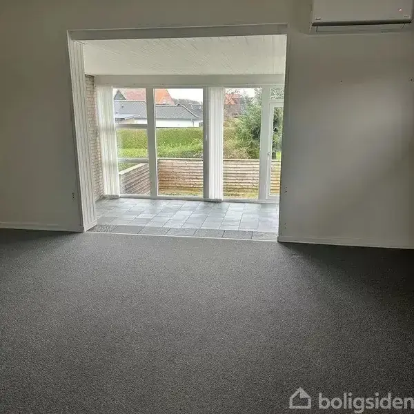 Gulvtæppe dækker stuegulv, der fører til flisebelagt udestue med store glasdøre. Udenfor er en have med hegn, og man kan skimte et hus i baggrunden.