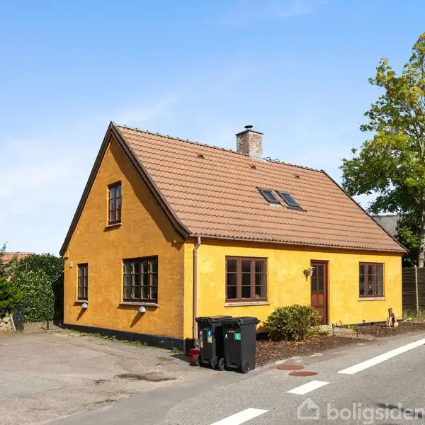 Et gulmalet hus med brunt tegltag står ved en vej. Ved siden af huset er en parkeret bil og nogle skraldespande. Omkring står træer og buske.