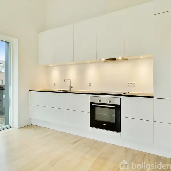 Et moderne, hvidt køkken med integreret ovn og kogeplade. Vasken er til venstre med en kromhane. Der er overskabe og lys under dem. Store vinduer fører ud til en balkon.