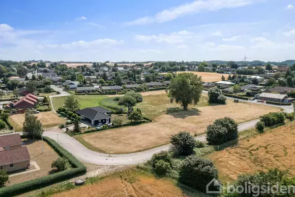 Landsbyhuse omgivet af grønt landskab og marker. En snoet vej fører gennem området, hvor træer står i rækker omkring en stor, åben mark. Vindmøller ses i horisonten.