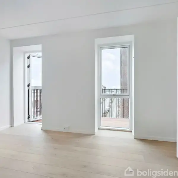 Et lyst rum med trægulv, to døre åbner ud til en balkon, hvor metalrækværk er synligt. Hvide vægge giver et minimalistisk præg. Ingen tekst i rummet.