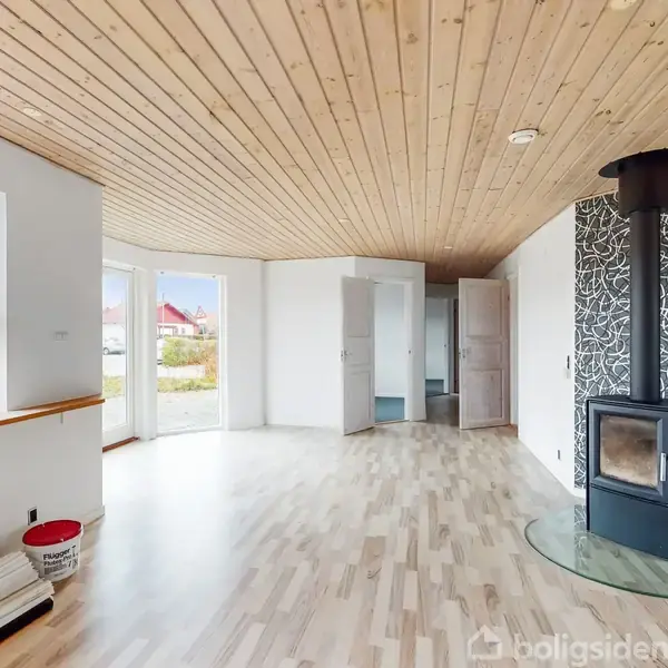 Stue med brændeovn på glasplade, lyst trægulv, og træloft. Flere vinduer giver udsigt til en have. Malingsbøtter og ruller er placeret på gulvet nær en væg.