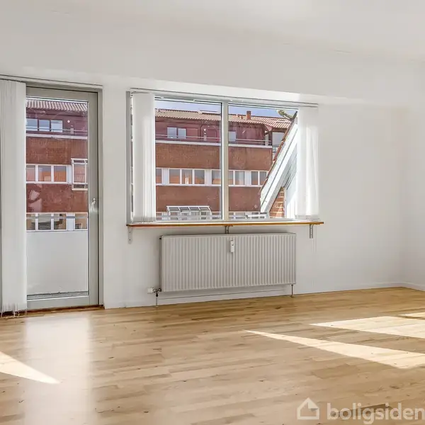 Et lyst rum med trægulv, store vinduer og glasdør, lader sollys strømme ind. Udsigt til røde murstensbygninger udenfor. Radiator under vinduet. Vægge og loft er hvide.