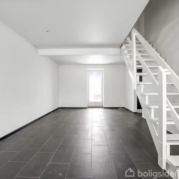 Trappe i en lys stue fører opad. Rummet har sorte klinker, hvide vægge og et stort vindue, der giver naturligt lys. Sparsom indretning bidrager til rummet.