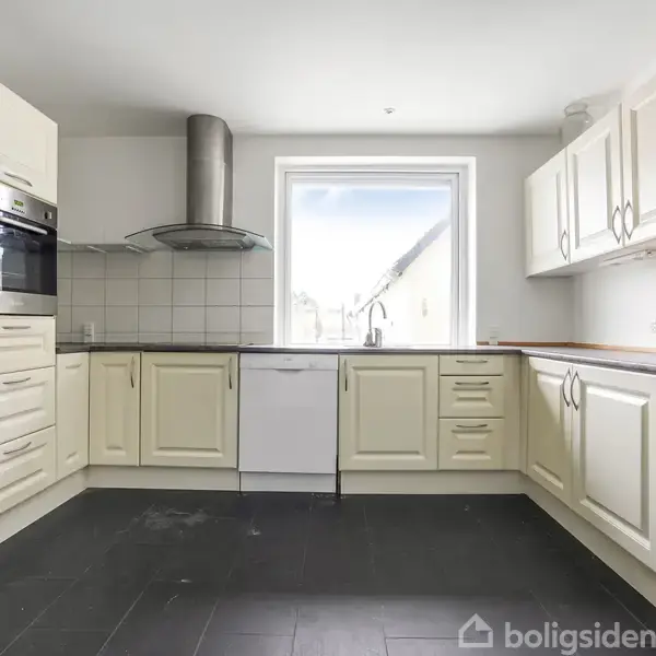 Køkken med lyse skabe og skuffer, et komfur og ovn indbygget i en sort væg, en opvaskemaskine under vinduet og en emhætte. Rummet har mørkt gulv.