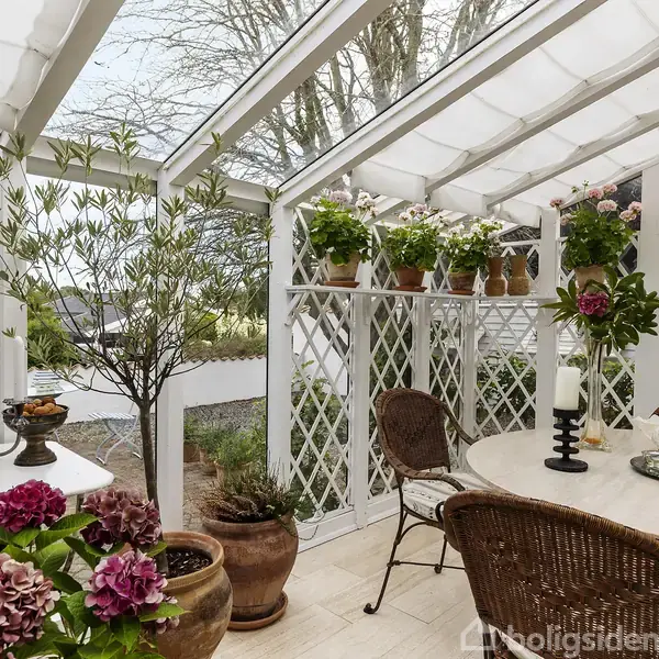 En overdækket terrasse møbleret med et rundt bord og stole. Planter i krukker og ophæng pryder rummet, der har hvidt espalier og glasloft. Ingen tekst til stede.