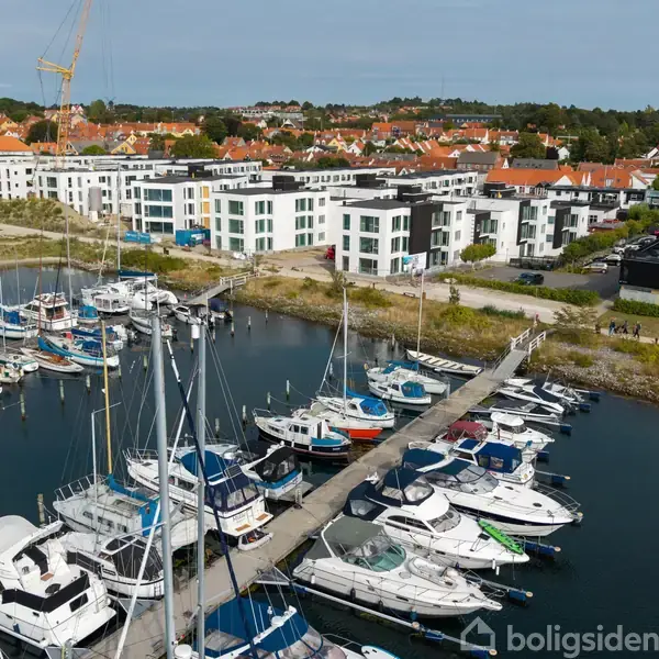 Både ligger for anker ved en moderne lystbådehavn med en rækkebyggeri i baggrunden og en kran tårnende over det. Bagved ses en by med røde hustage.